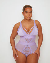 Model wearing the Alicia Fuller Bust Curvy Girl Bodysuit in Soft Lavender, featuring a plunging neckline with decorative strap detailing, floral lace cups, sheer mesh panels, and a high-waisted silhouette designed to flatter and support fuller busts.