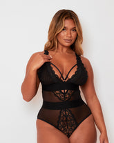 Model wearing the Alicia Bodysuit in Black by Tutti Rouge, featuring sheer mesh panels, floral lace detailing, a plunging neckline with strappy accents, and contouring waistband panels for a flattering silhouette.