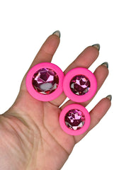 Pink circularbutt plugs with gemstone-like centers held in a hand against a white background