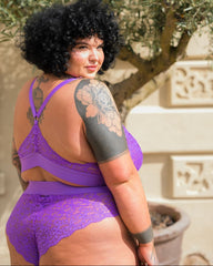 Curvy model wearing the Hallie lace bralette and high-waisted brief set in Passion Purple by Tutti Rouge, posing outdoors with back detail visible and floral lace design showcased.