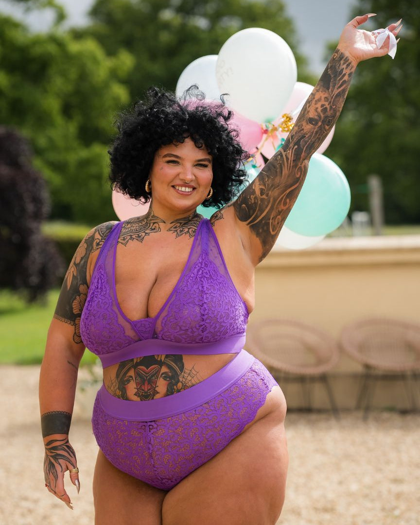 Smiling tattooed curve model wearing the Hallie Passion Purple lace bralette and matching briefs from Tutti Rouge, standing outdoors with balloons in the background.